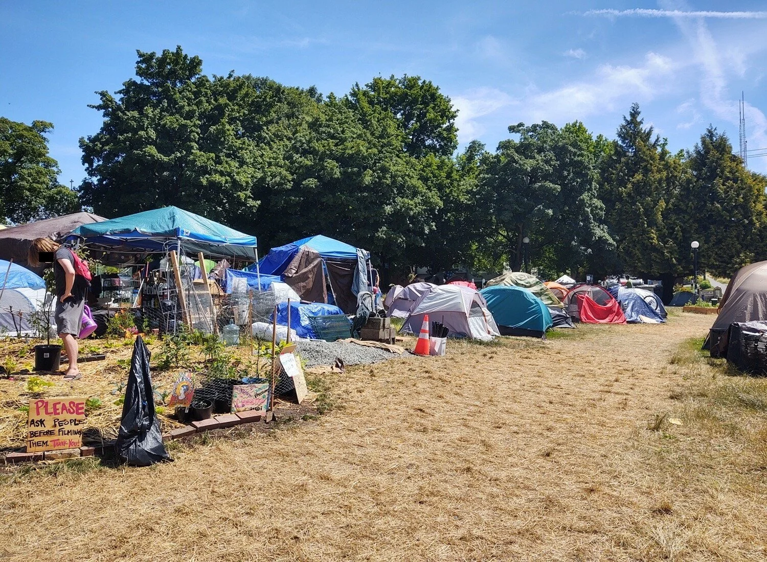 A person waters the CHOP garden with a purple watering can near garden supplies and tents, under lush, green trees.