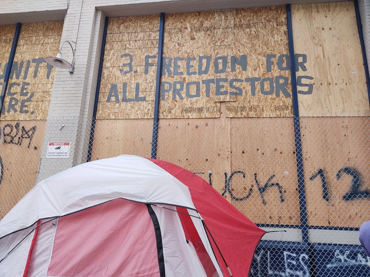 “3. Freedom for all protesters” is written on a boarded-up window. A red and white tent is in the foreground.