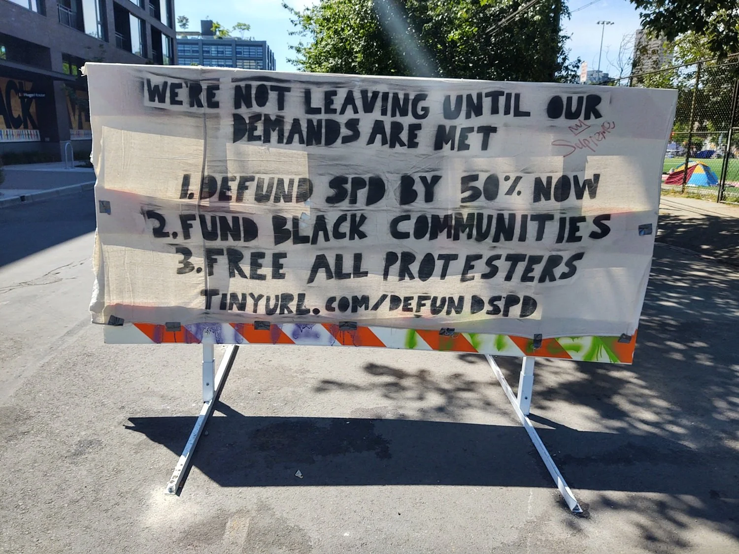 Banner attached to orange and white traffic barricade insists protesters aren’t leaving, lists three demands, and a link.