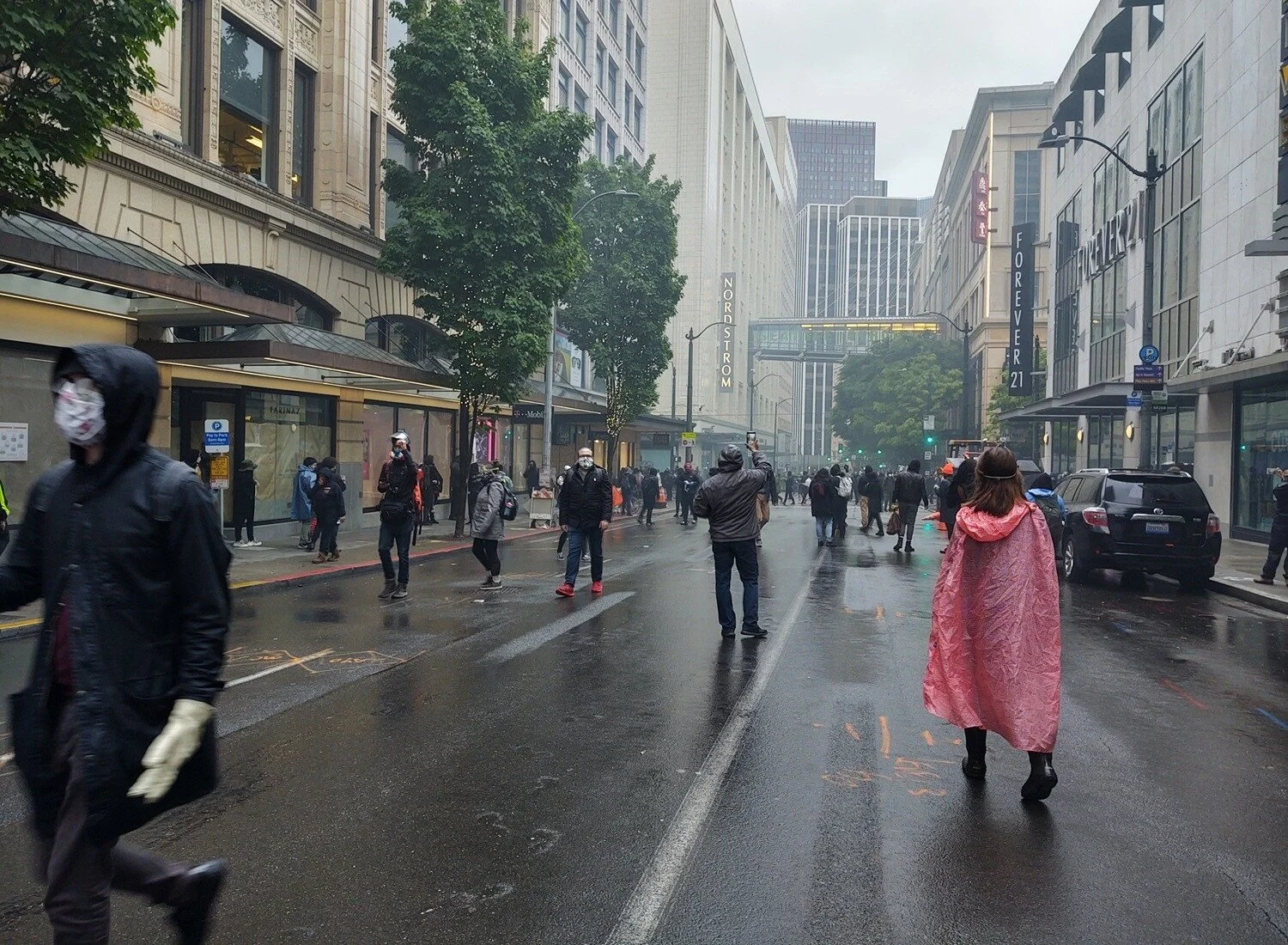 Protesters walk on a wet street in a commercial area wearing ponchos, coats, masks, and gloves.