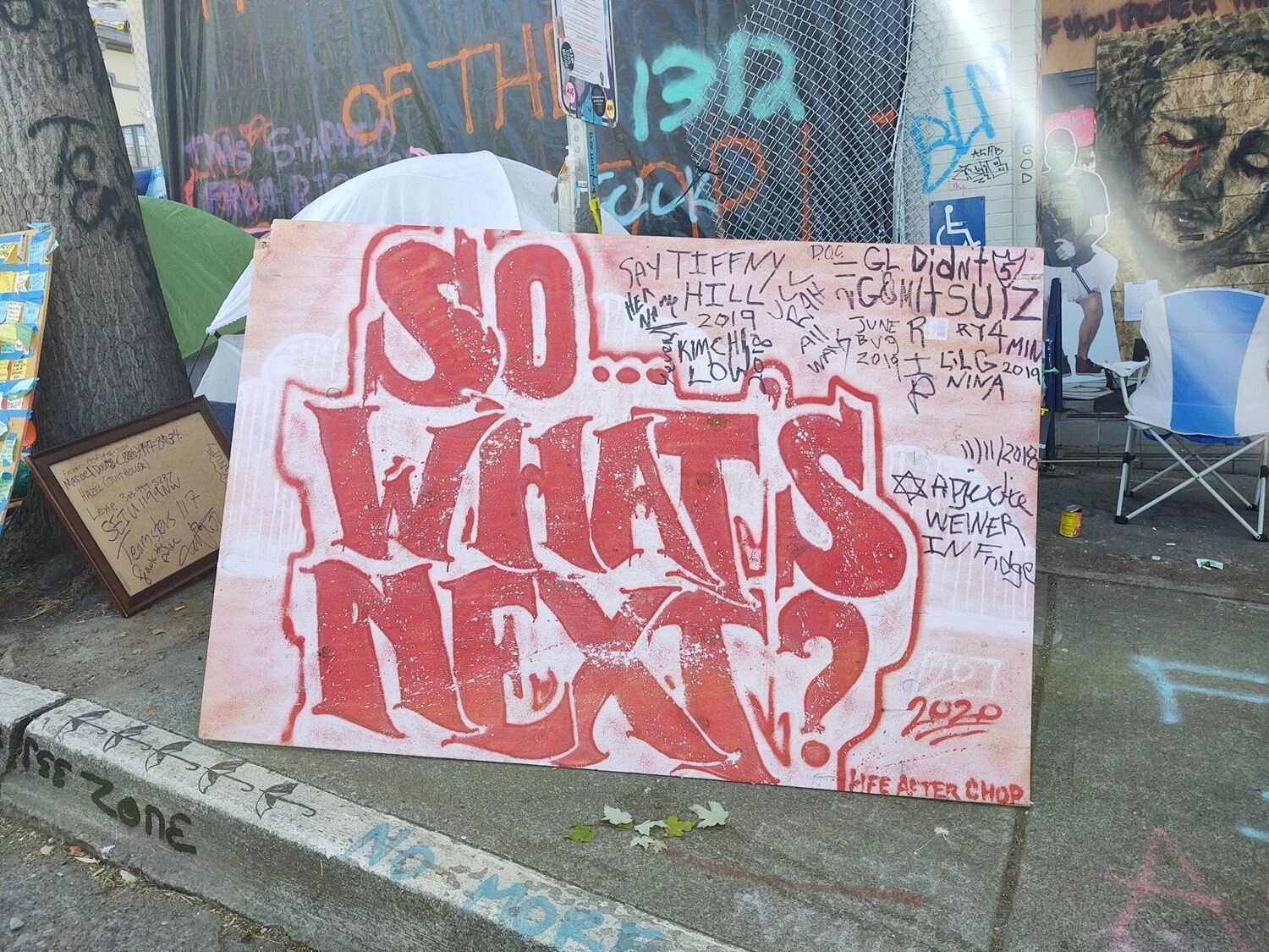 Red text on a rectangular piece of plywood, leaning against a street sign pole outside the East Precinct.