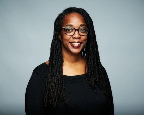 Tarika Powell poses for a professional photo wearing a black shirt and glasses, with long dreadlocks, in front of a blue background.