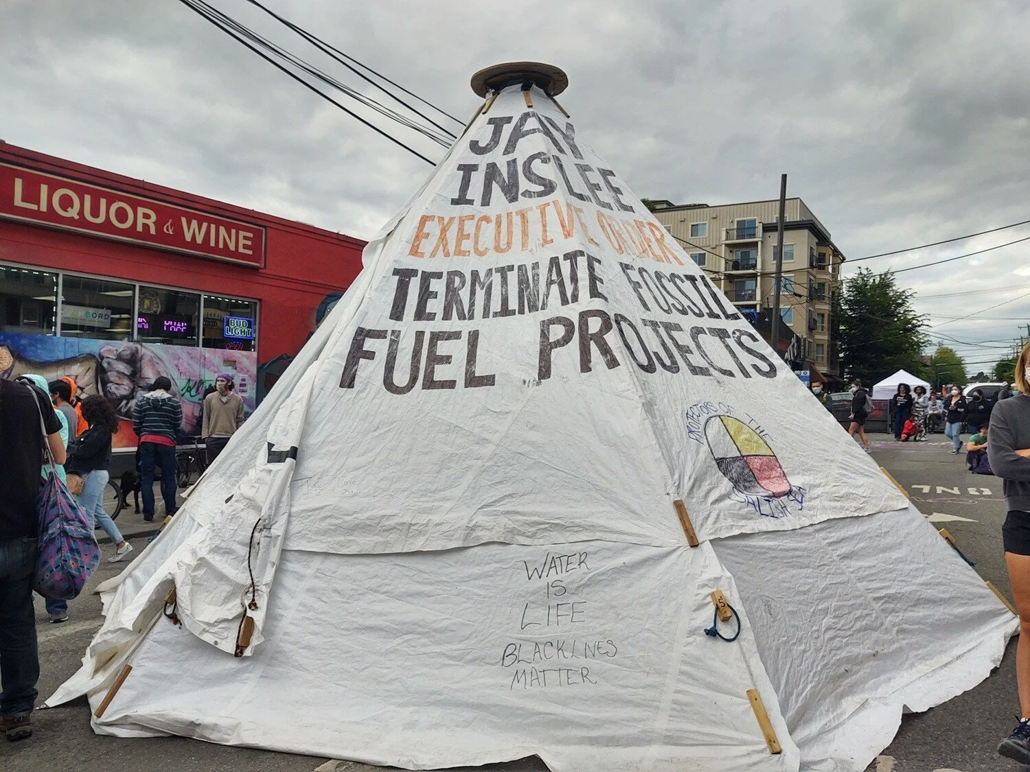 White tipi with block letters in front of NW Liquor and Wine. “Jay Inslee. Executive Order. Terminate fossil fuel projects.”