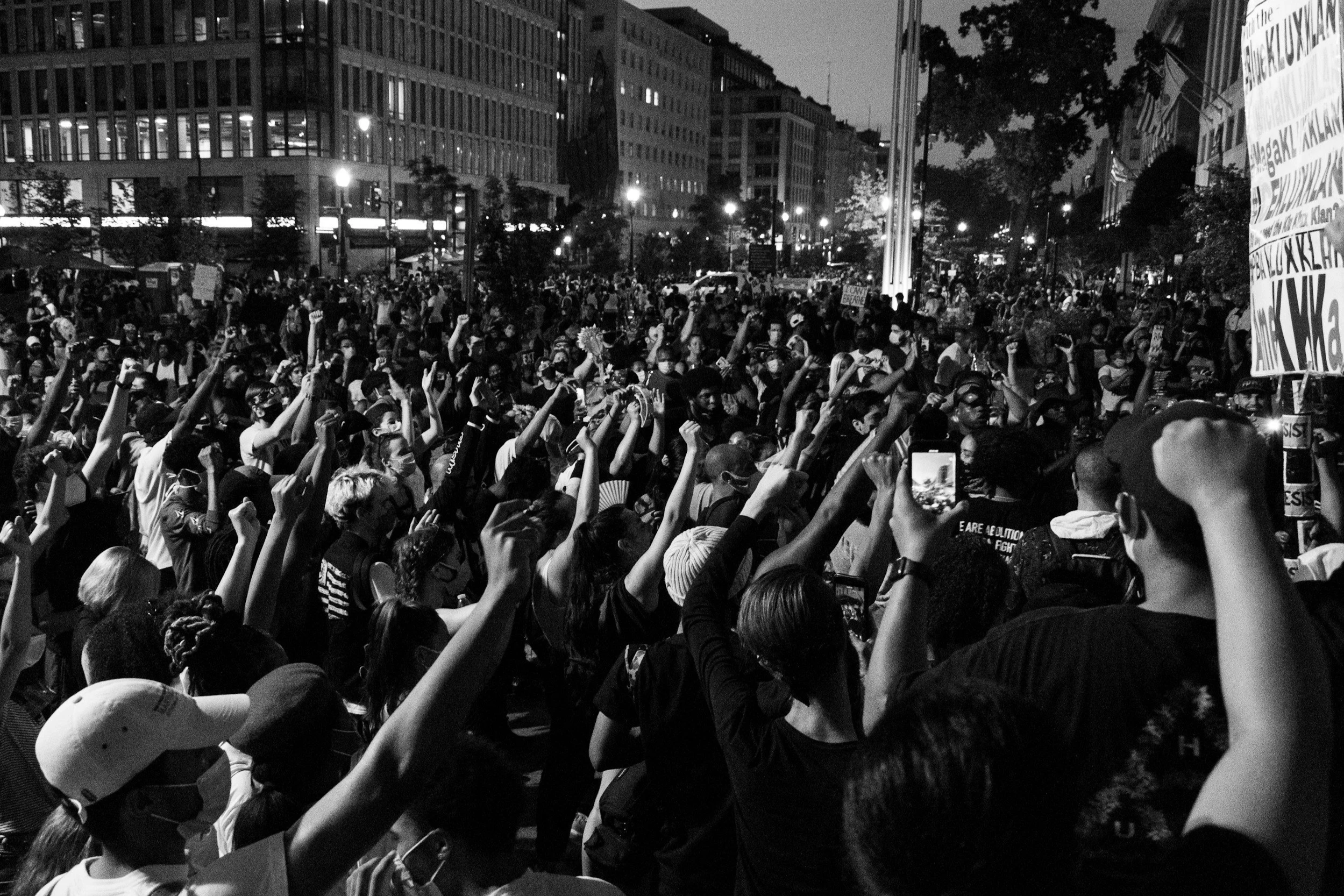 A large crowd of people gathered outdoors at night, with raised fists, holding signs. Tall buildings and streetlights are visible in the background.