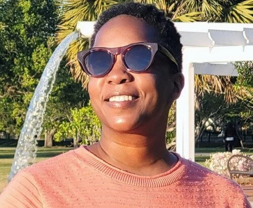 Writer Tarika Powell, TarikaWrites, is pictured in an outdoor park wearing an orange shirt, with short hair and sunglasses.