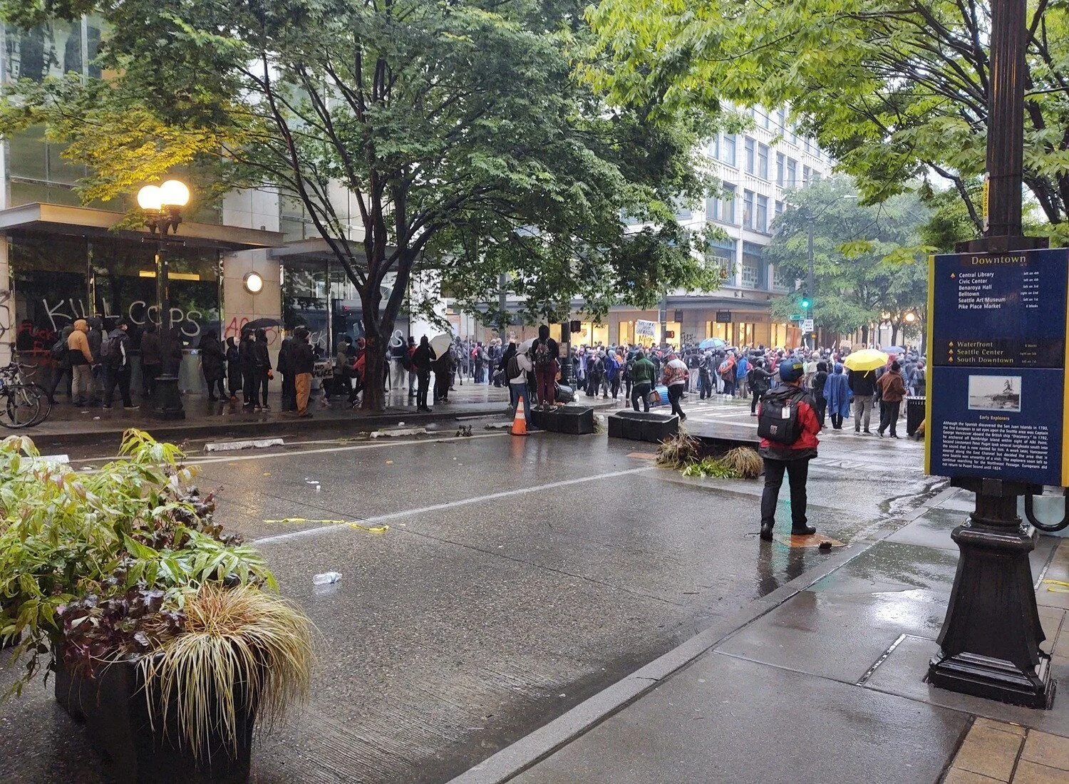 Hundreds of protesters stand in the street or huddle under store awnings, some holding umbrellas.