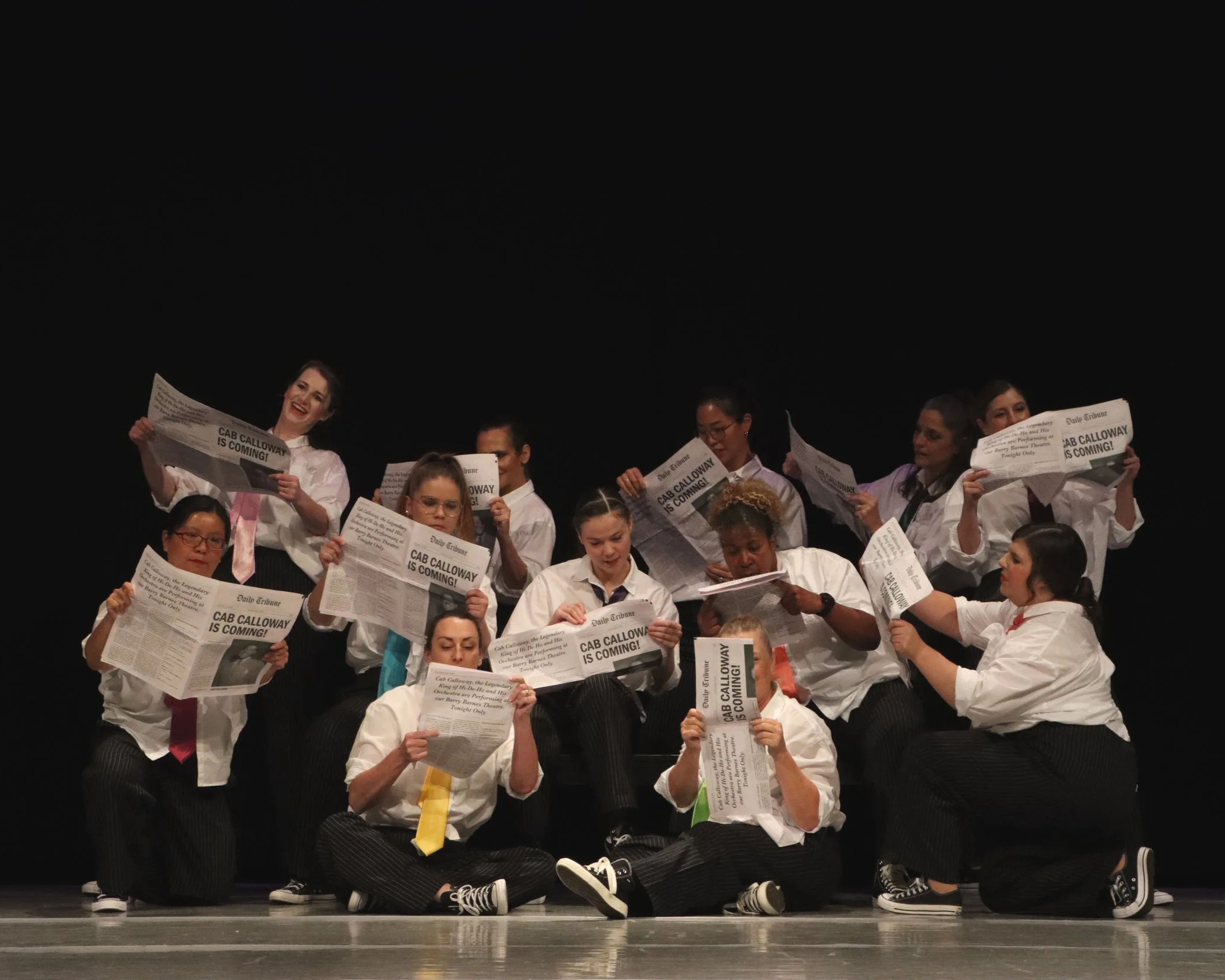 Group of people dressed in white shirts and black pants sitting and kneeling on stage, reading newspapers with headlines about Cab Calloway, against a black background.