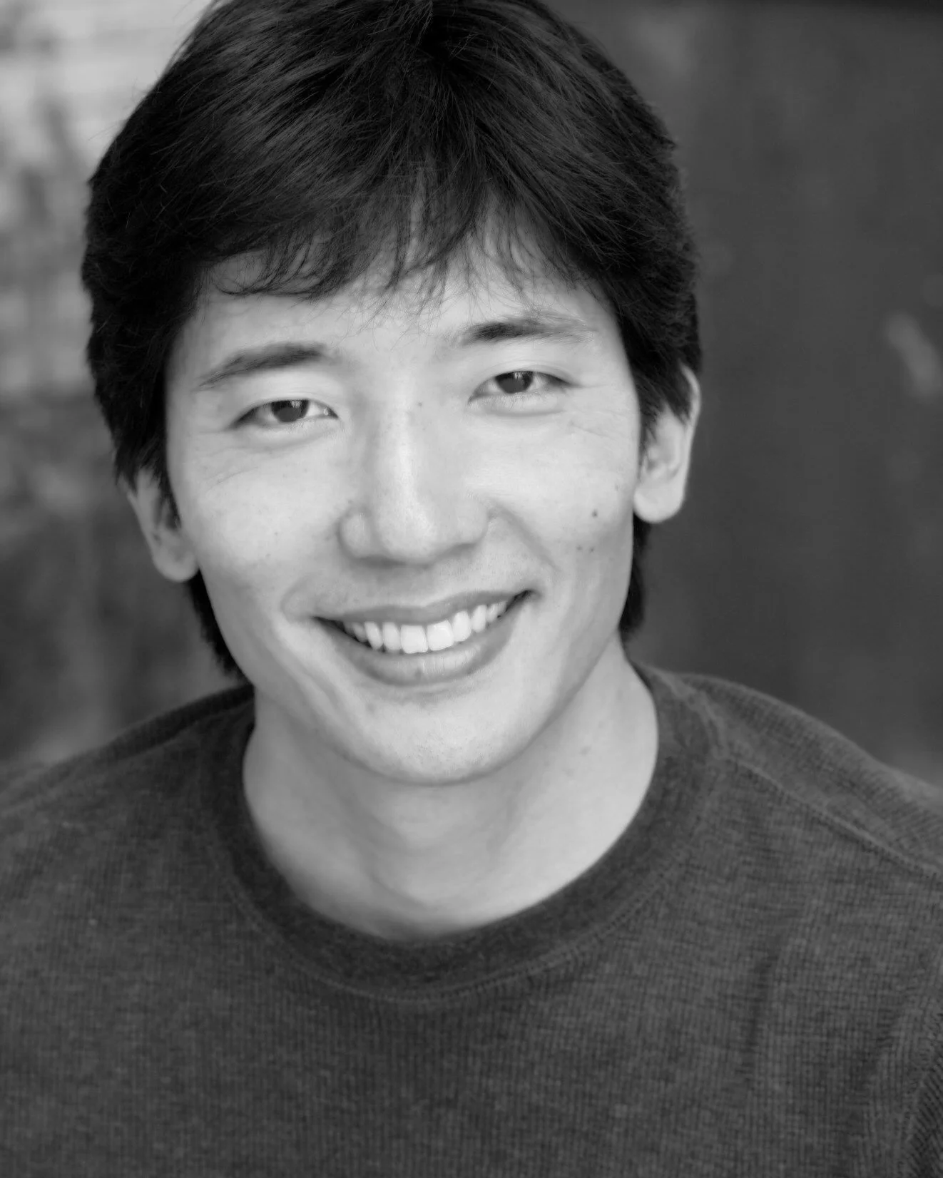 Black and white photo of a smiling man with short, dark hair wearing a dark shirt, looking directly at the camera.