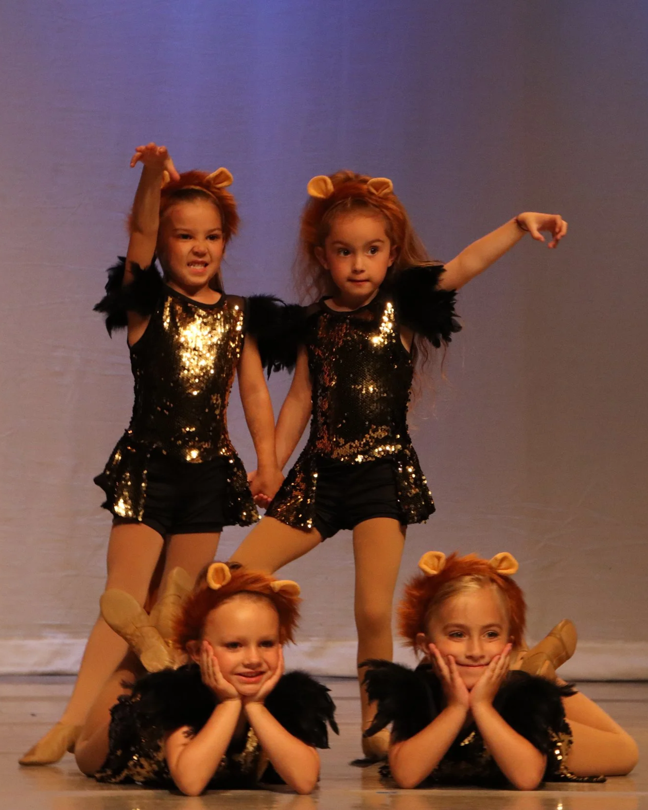 Group of young children dressed in lion costumes performing a dance on stage, with two girls standing in the back and two lying down in front, holding their faces with hands, against a plain background.