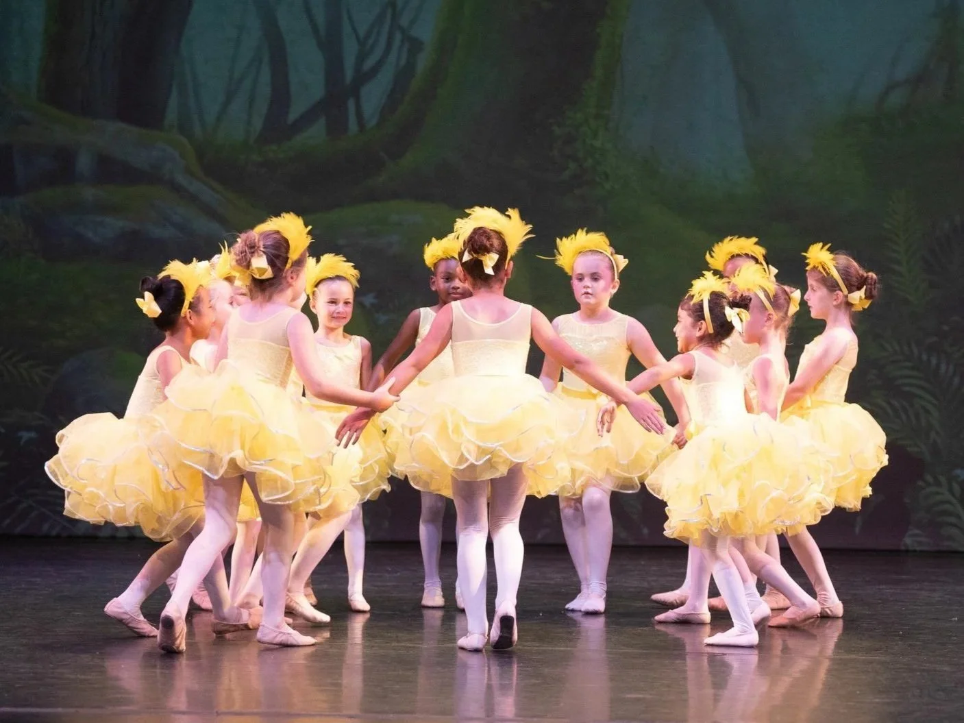 Group of young girls dancing in yellow tutus and headbands on stage with a forest backdrop.