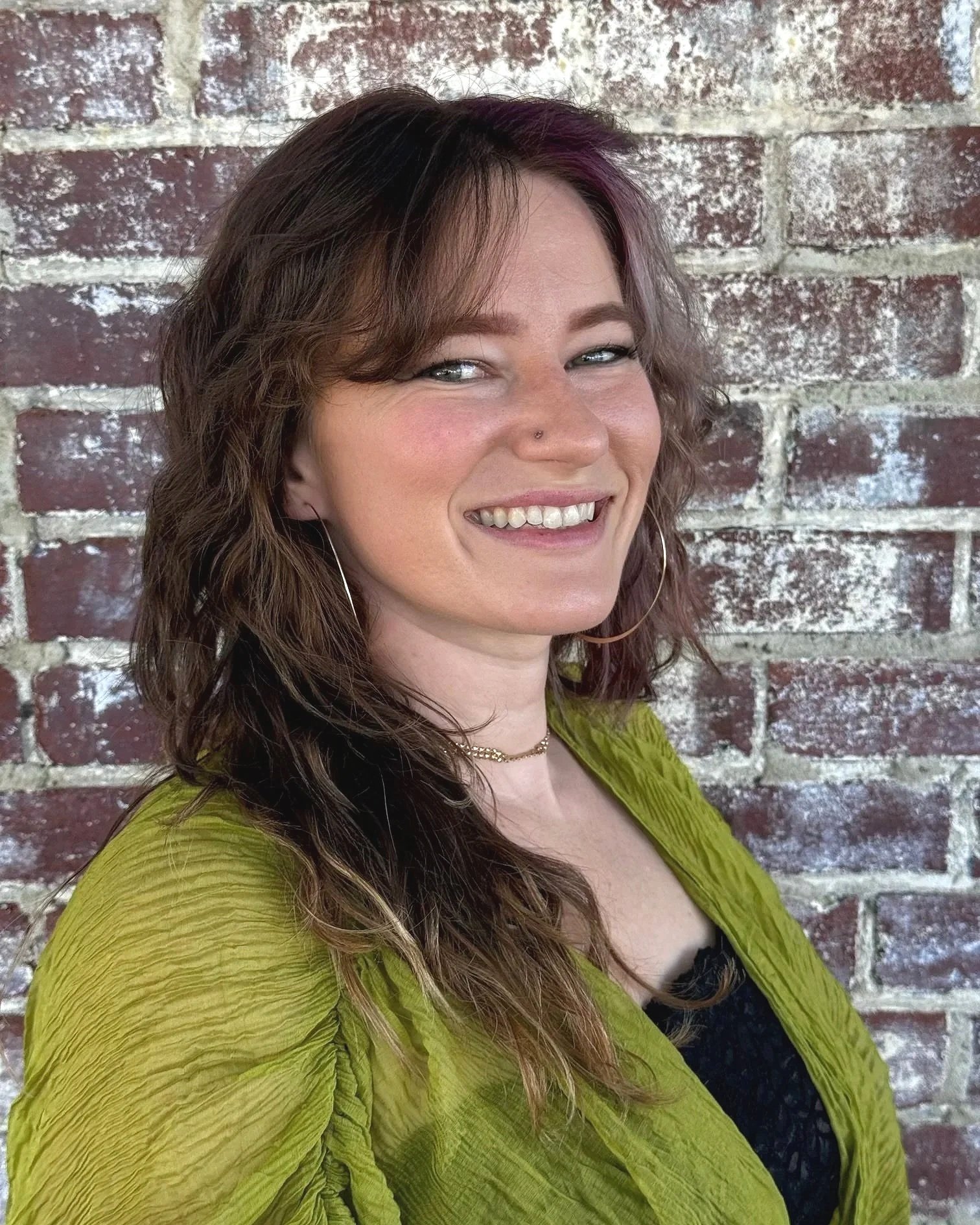 A woman with curly brown hair and a nose piercing smiling in front of a brick wall, wearing a green top and hoop earrings.