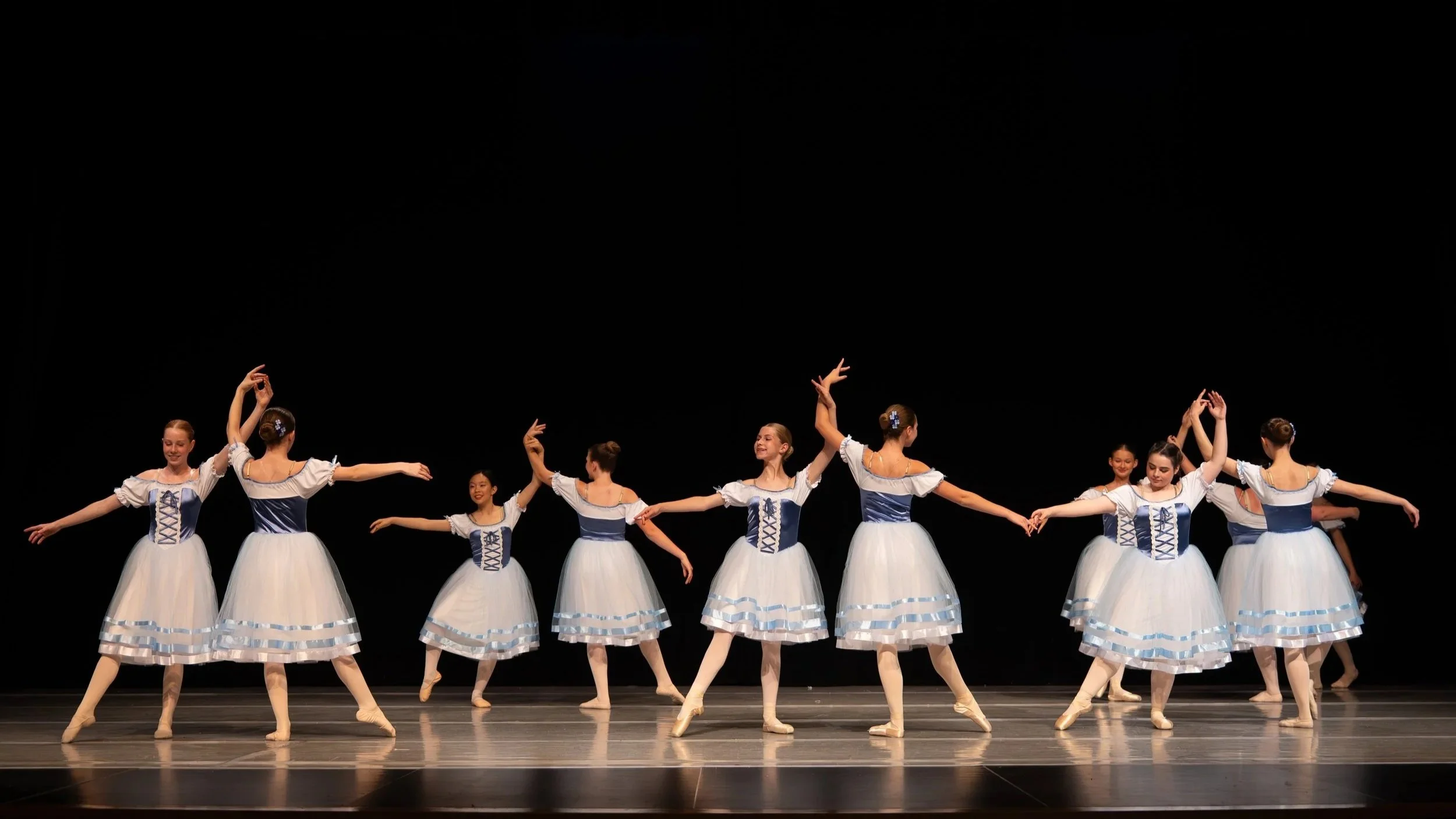 Group of young ballet dancers performing on stage in white and blue costumes, holding hands in a line.