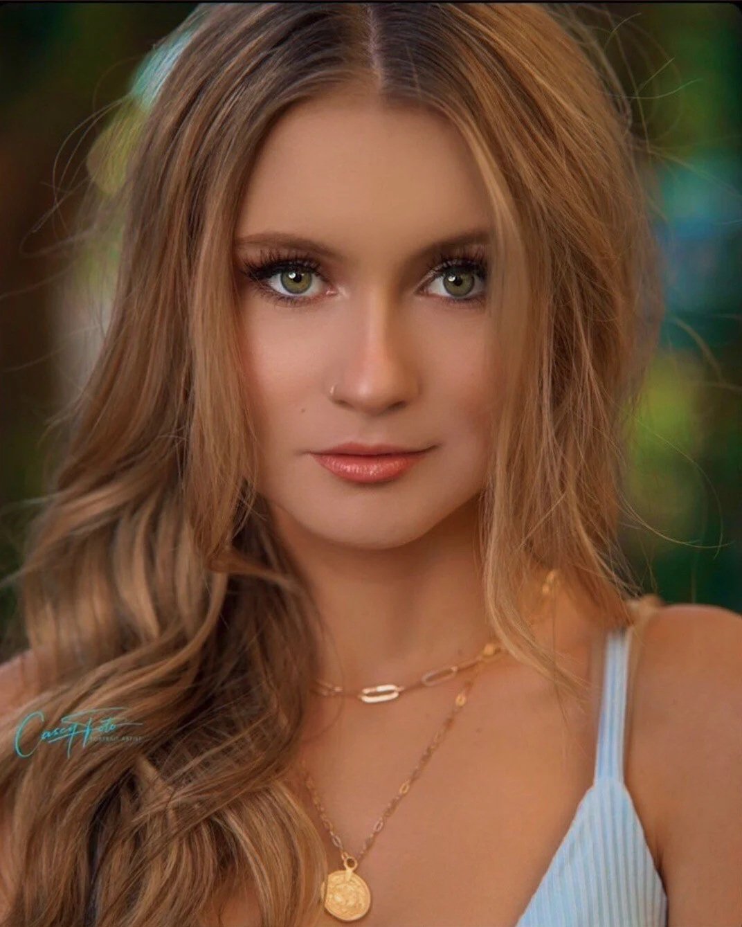 Close-up of a young woman with long, wavy, light brown hair and green eyes, wearing a white striped top and layered gold necklaces, outdoors with blurred greenery in the background.