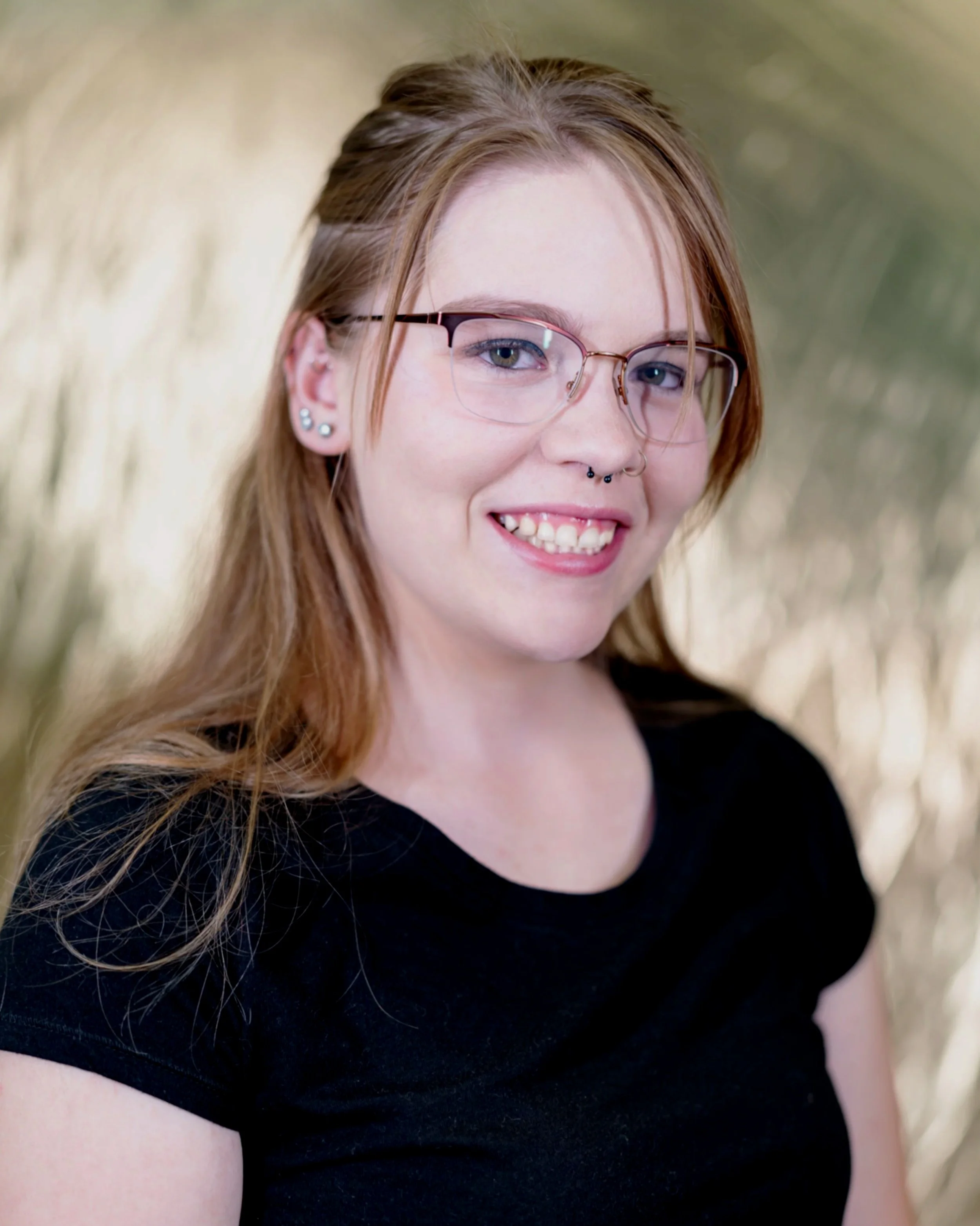 Close-up of a young woman with long red hair, glasses, multiple earrings, and a septum piercing, smiling while wearing a black top.