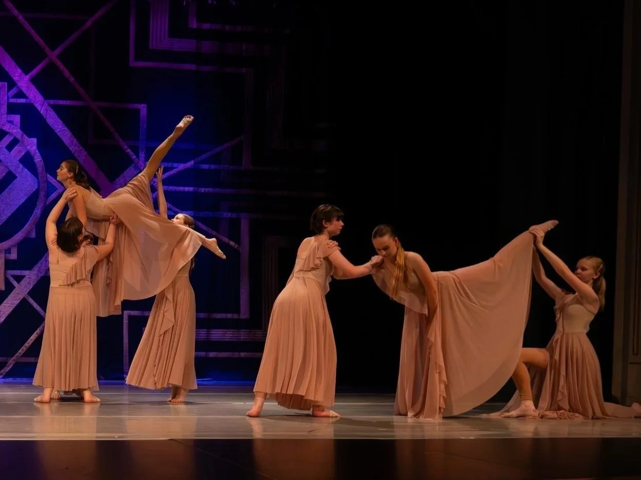 Six young women dressed in peach-colored dresses perform a contemporary dance on stage with a dark background and geometric purple lighting design.