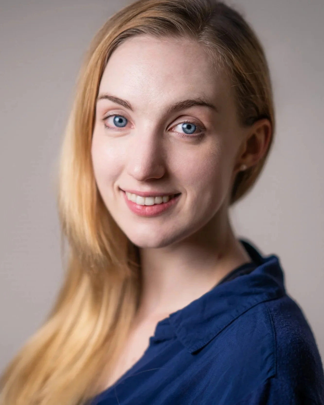 Close-up of a young woman with long red hair, blue eyes, and a bright smile, wearing a dark blue top, against a plain background.