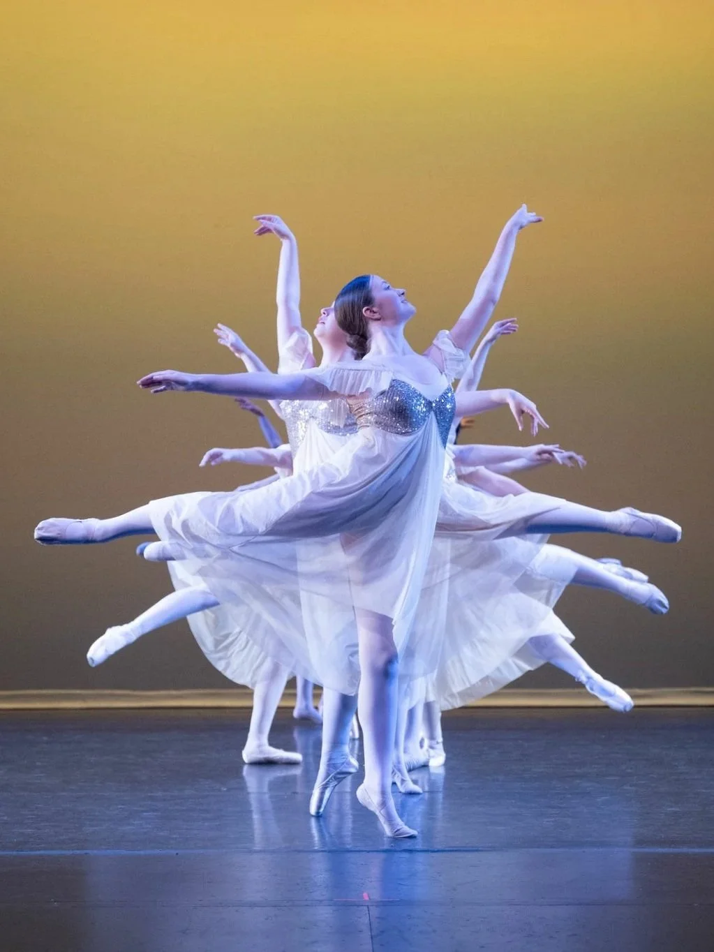 Ballet dancers performing on stage with a yellow gradient background, dressed in white tutus and pointe shoes, in a synchronized pose.