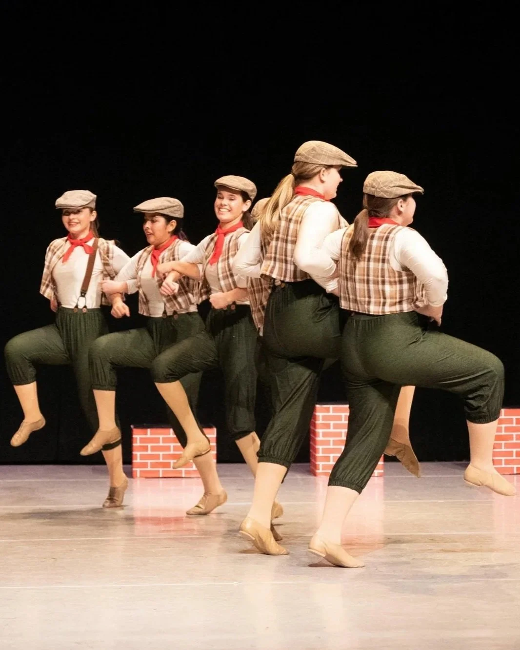 A group of six young girls dressed in 1920s-style costumes performing a dance line on stage with a black background and brick props.