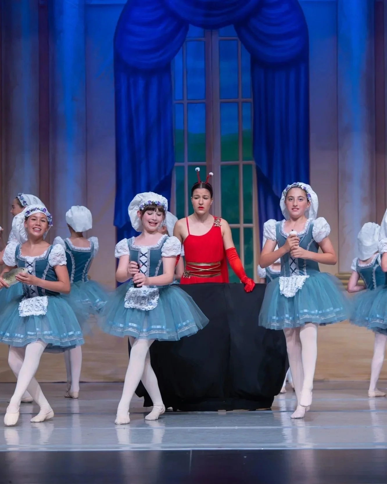 Children in ballet costumes performing on stage with a woman in a red dress and antenna headband. The stage has a blue curtain backdrop with a window scene.