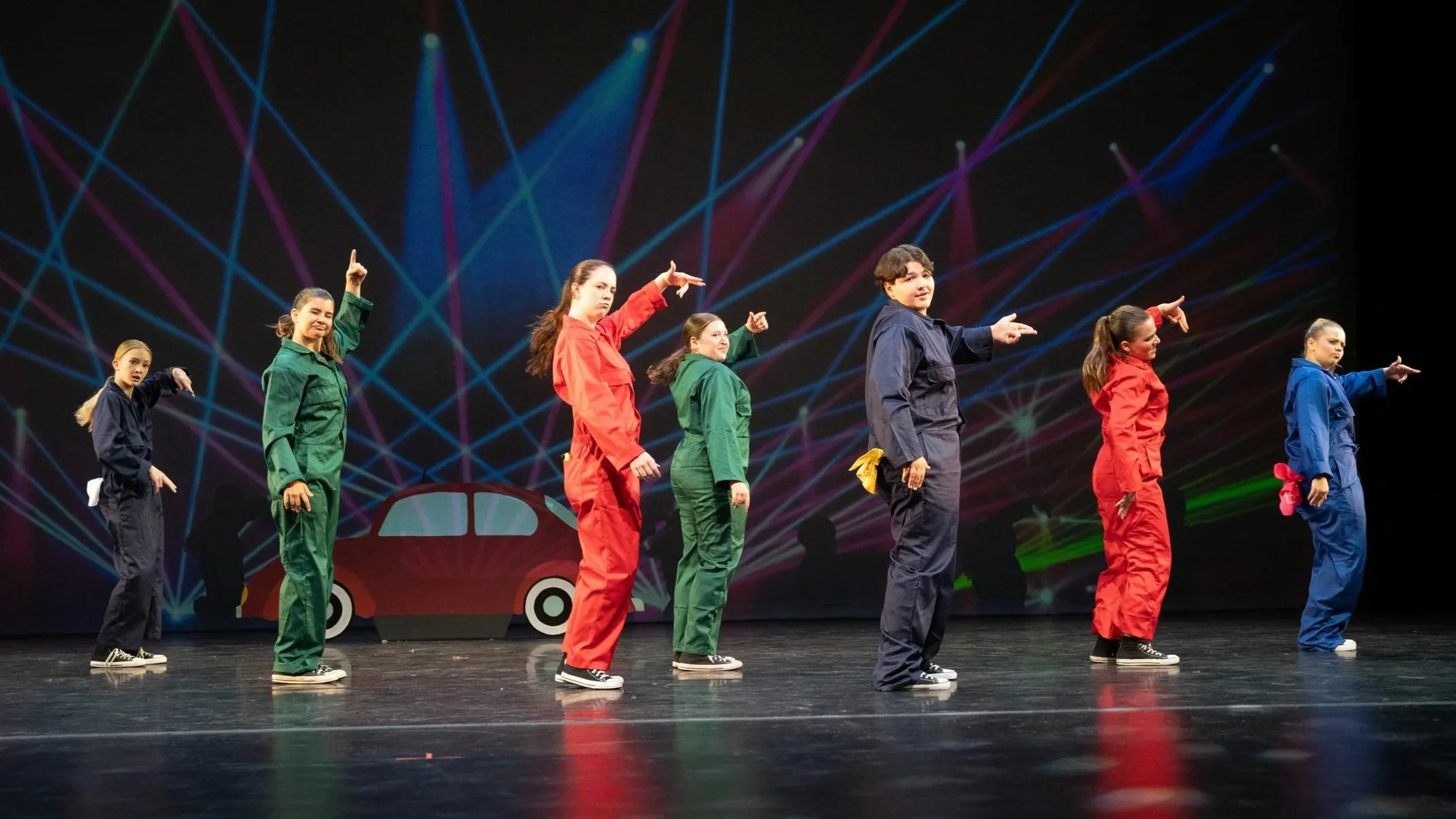 Group of young performers in colorful coveralls on stage with a background of laser lights and a car illustration.