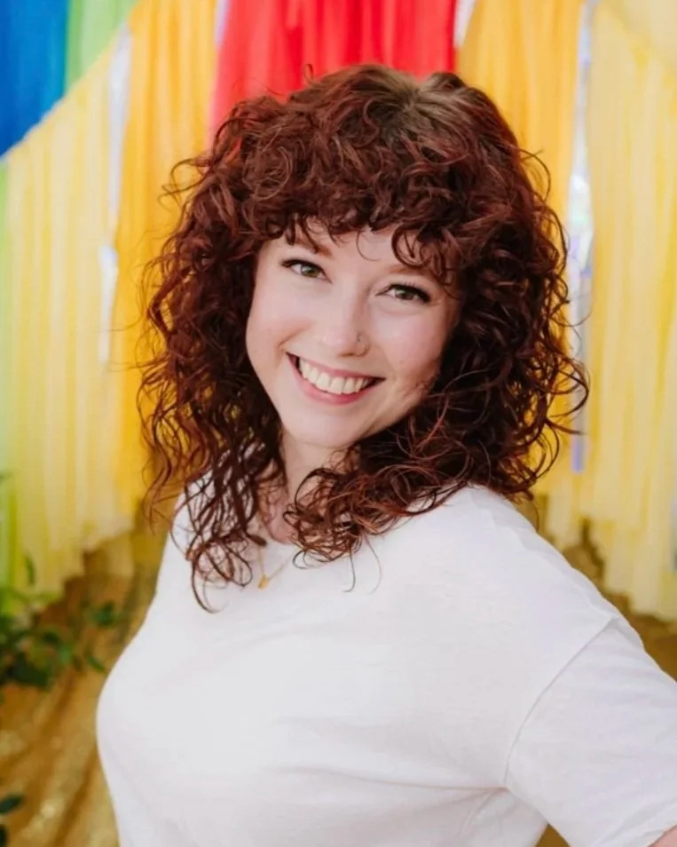 A woman with curly red hair and a white shirt smiling, standing in front of multicolored ribbons or fabric strips.