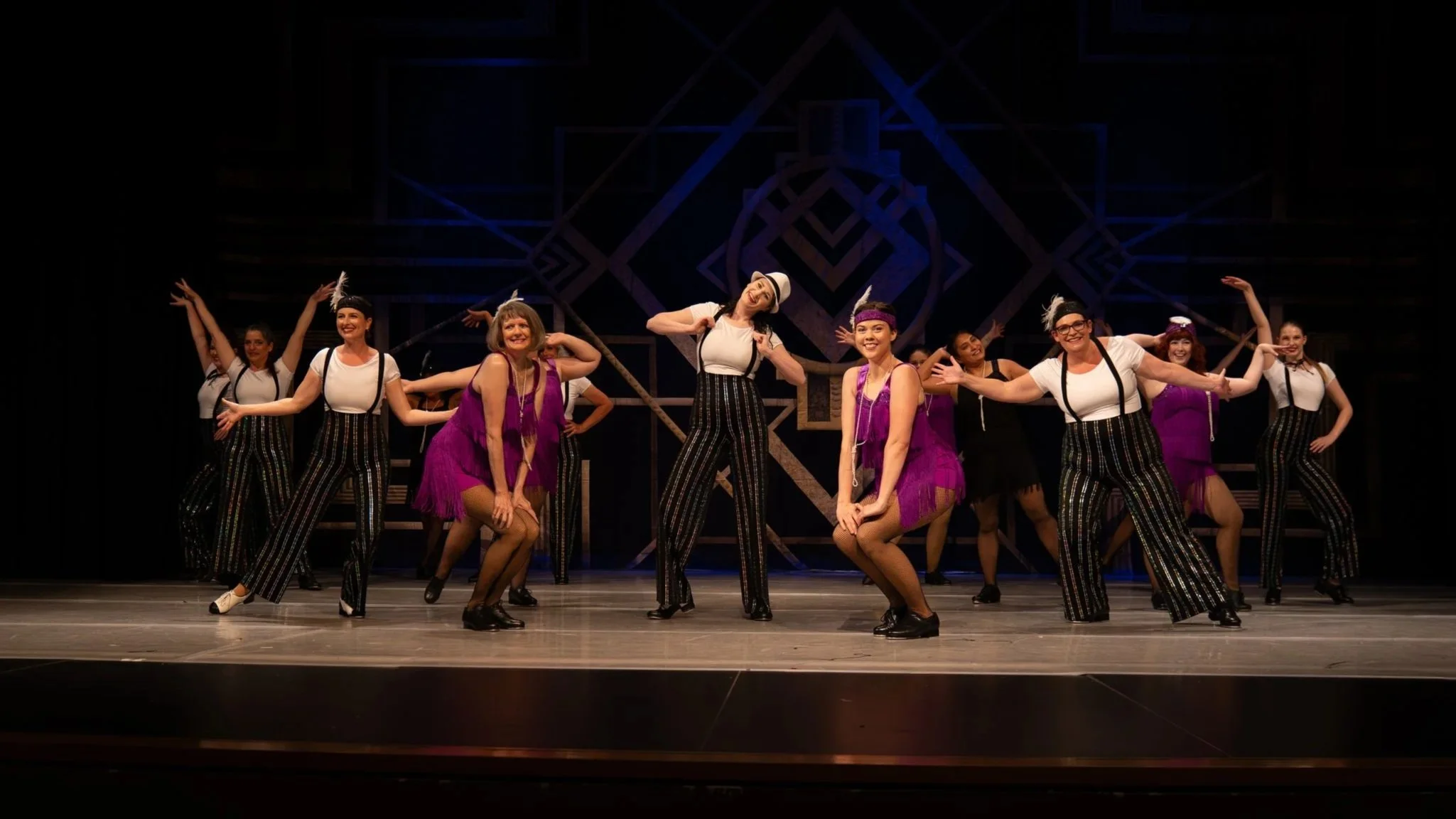 Group of women performers on stage, some in black and white striped pants, others in purple dresses, dancing with poses and smiles, stage with geometric backdrop and dark lighting.
