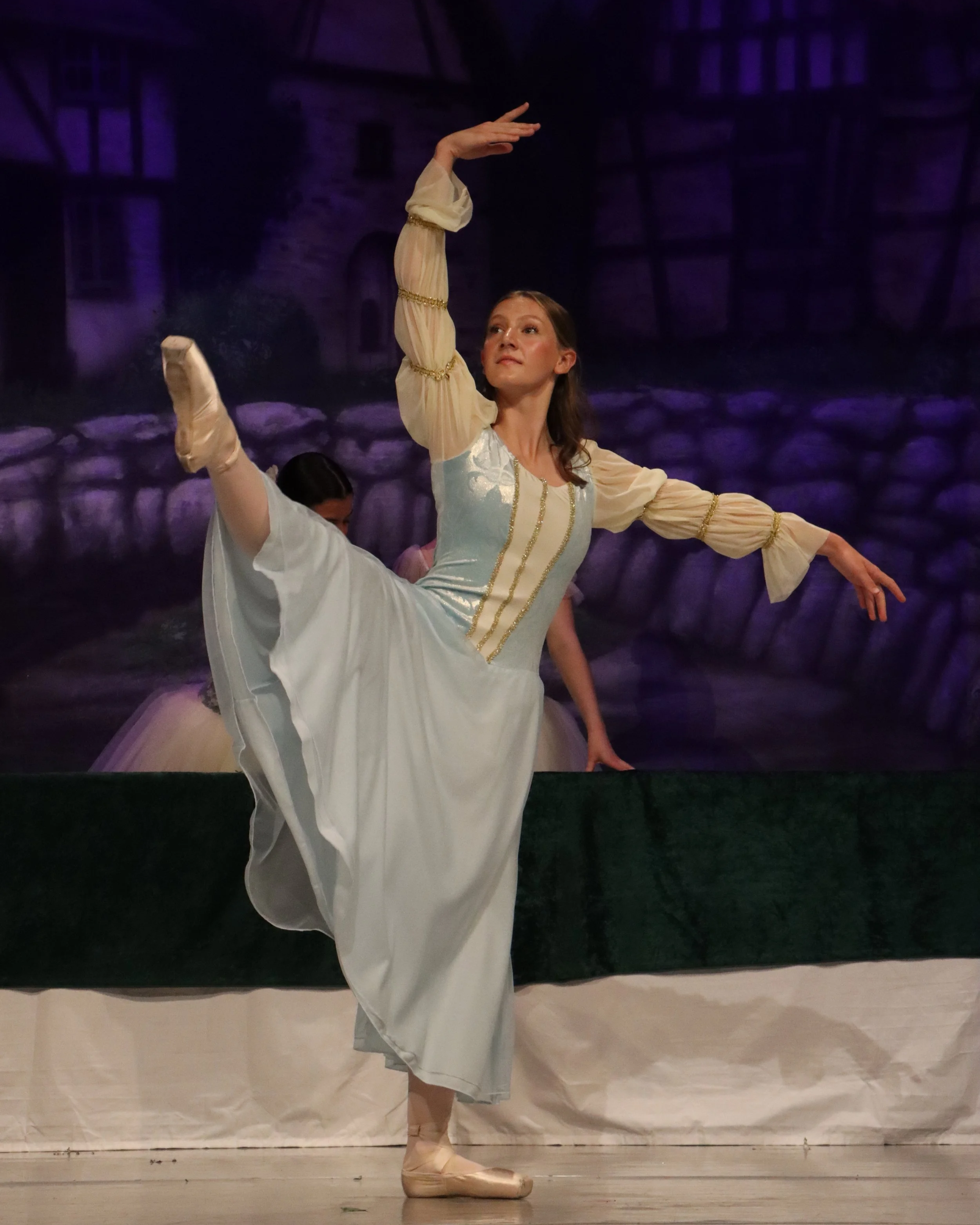 A ballerina in a classical pose on stage, wearing a white tutu and pointe shoes, with a dark theatrical background.