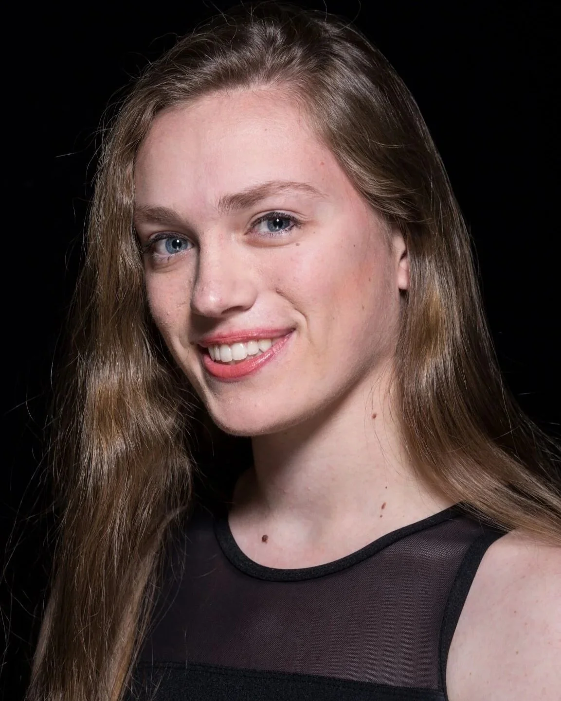 A young woman with long, wavy brown hair and blue eyes smiling against a black background.