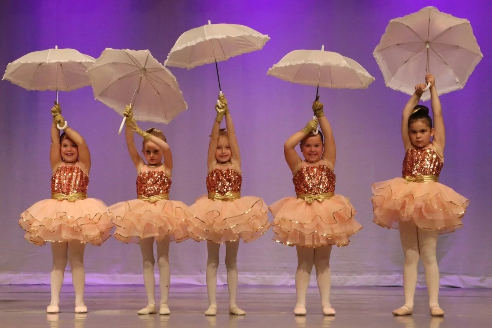 Five young girls in ballet costumes holding umbrellas on stage with a purple background.