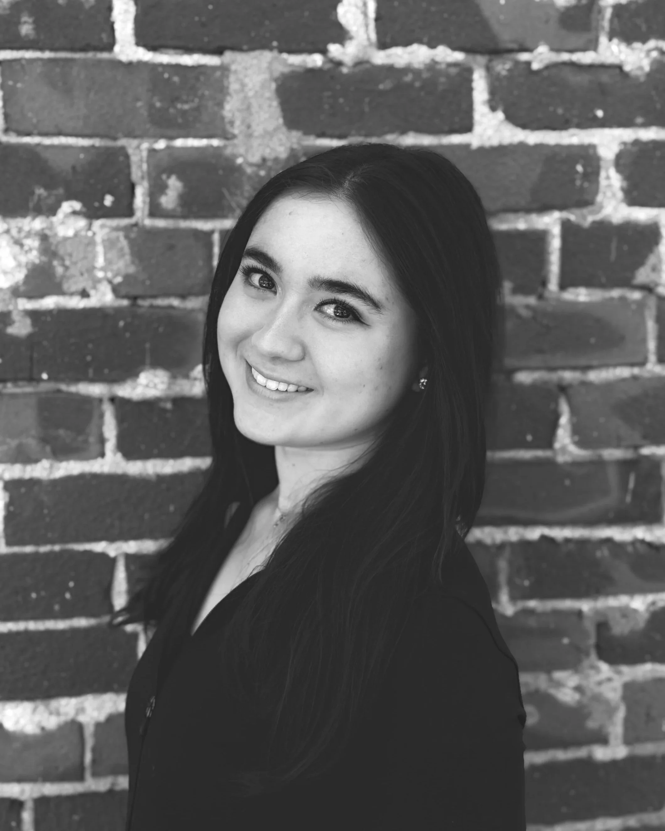A black and white photograph of a young woman with long dark hair smiling, standing against a brick wall background.