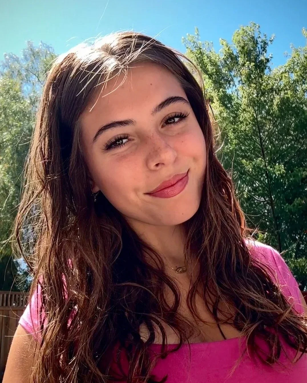 A young woman with long brown hair and a pink top, smiling outdoors with green trees and a blue sky in the background.