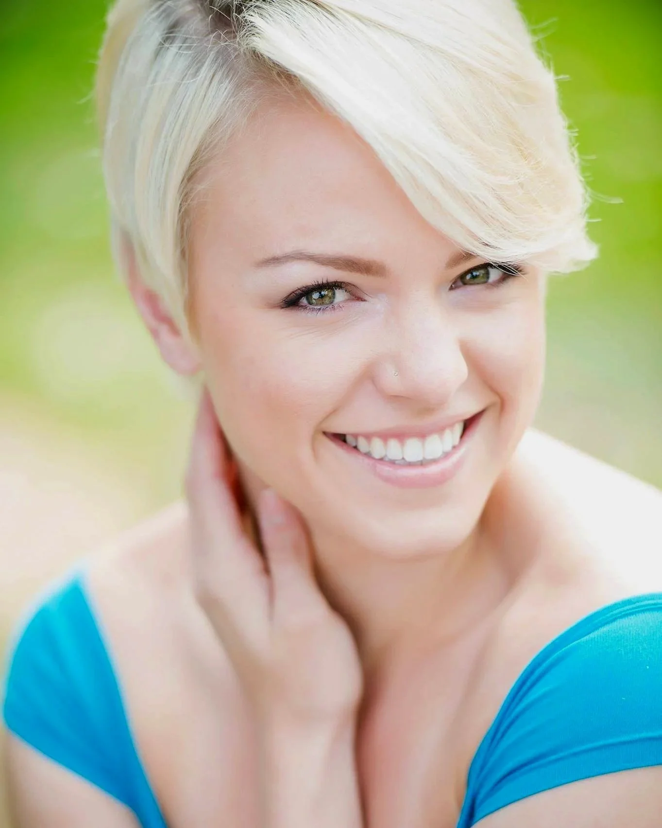 Close-up of a smiling woman with short blonde hair, green eyes, wearing a blue top, against a blurred green background.