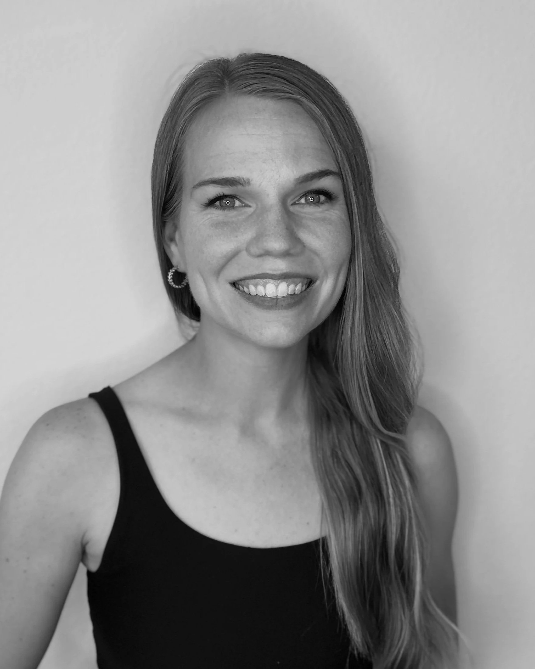 A woman smiling at the camera, wearing a black tank top and hoop earrings, with long hair, against a plain background.