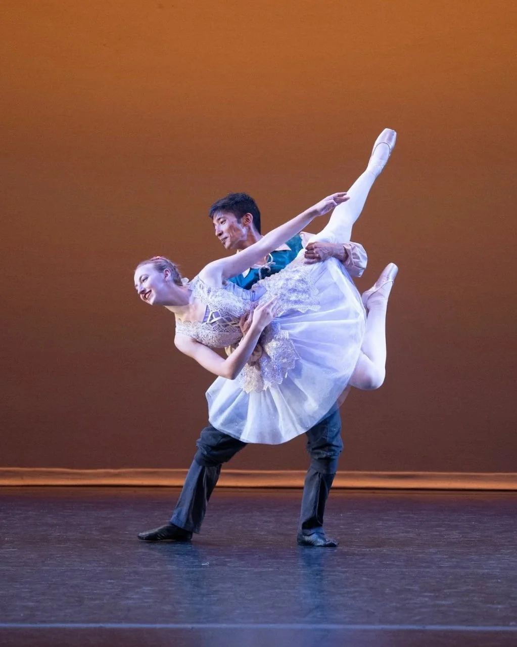 Ballet dancers performing on stage, with one dancer in a white tutu and another in a dark shirt and pants, against a plain brown background.