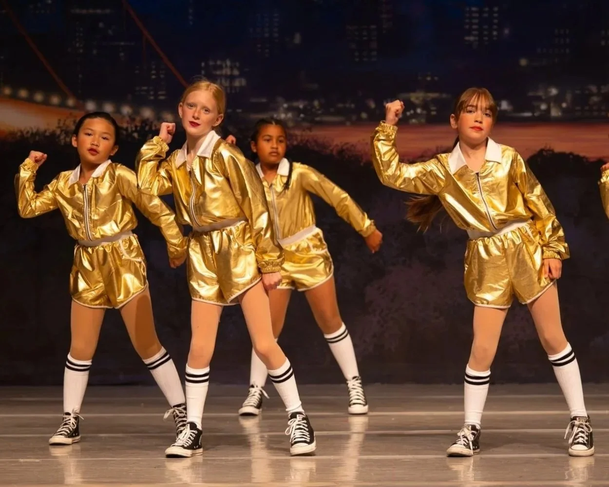 Young girls in gold jackets and shorts performing a dance on stage with city skyline backdrop.