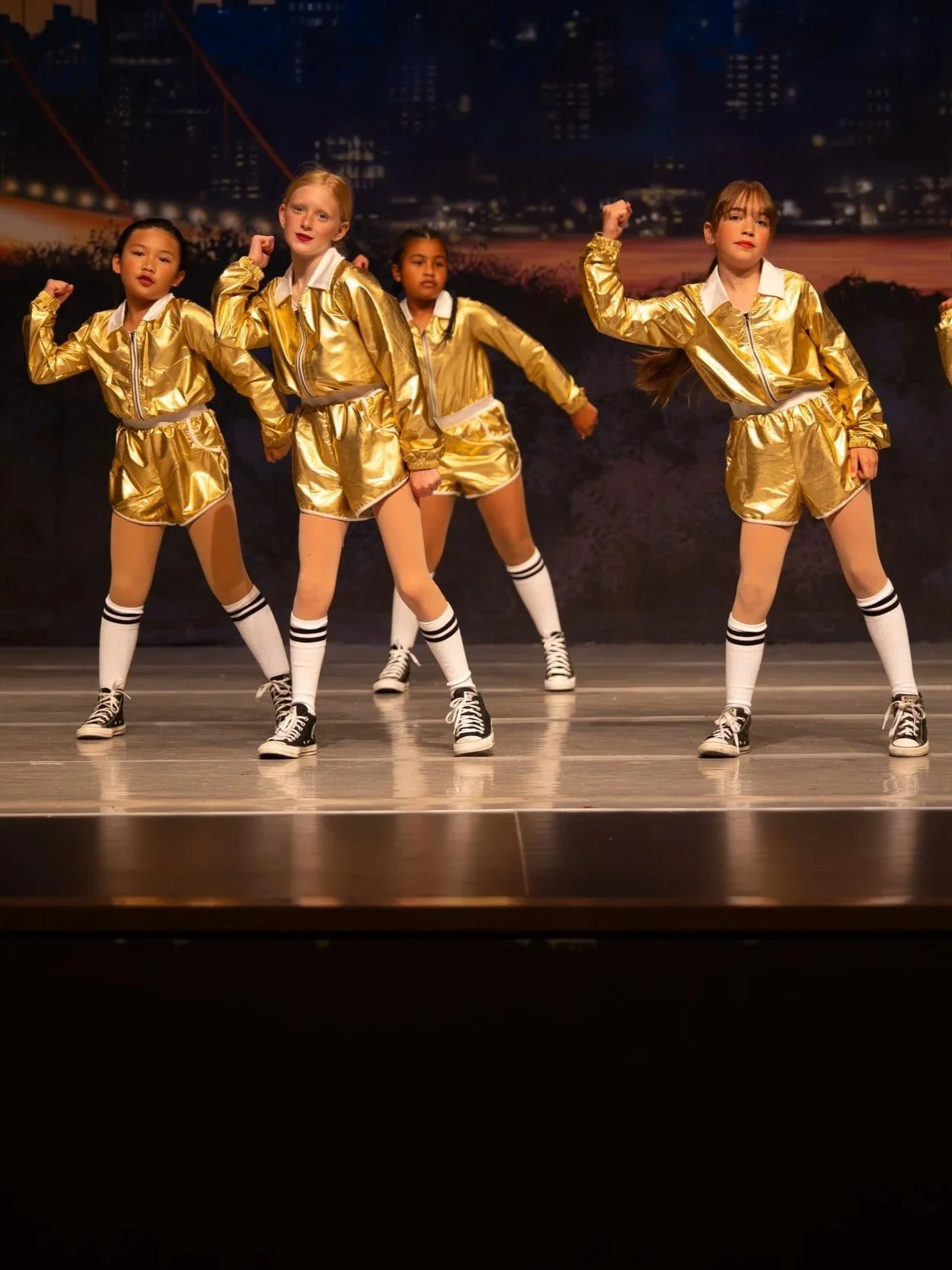 Group of young girls dressed in shiny gold jackets, shorts, and knee-high socks performing a dance on stage with a cityscape background.
