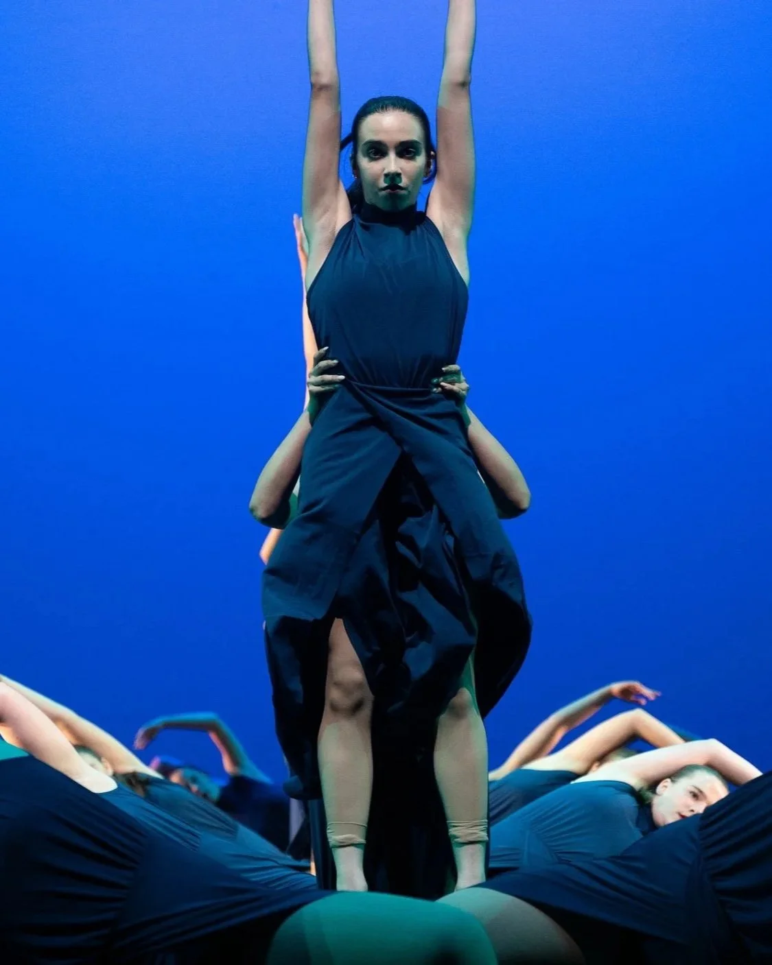 A group of performers creating an acrobatic formation on stage with a blue background, featuring a woman standing upright with arms raised, supported by others lying on the ground.