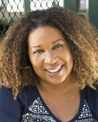 A woman with curly hair smiling outdoors in front of a black metal fence.