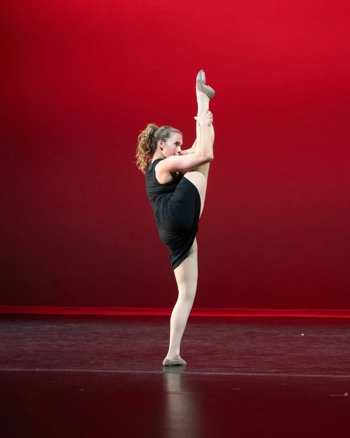 A woman in black dance attire performing a standing split with a red background.