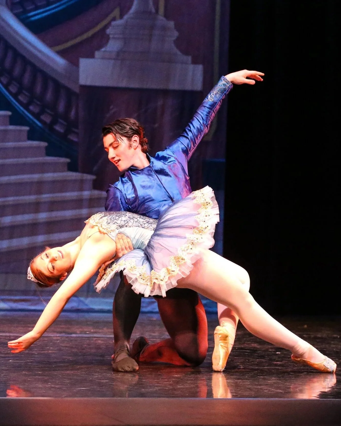 Two ballet dancers performing on stage; the male dancer is kneeling while holding the female dancer, who is leaning back in a ballet pose, wearing a tutu and tiara, with a staircase backdrop in the background.