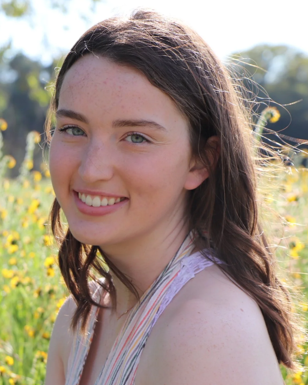 A young woman with long brown hair and blue eyes smiling outdoors in a field of yellow flowers.