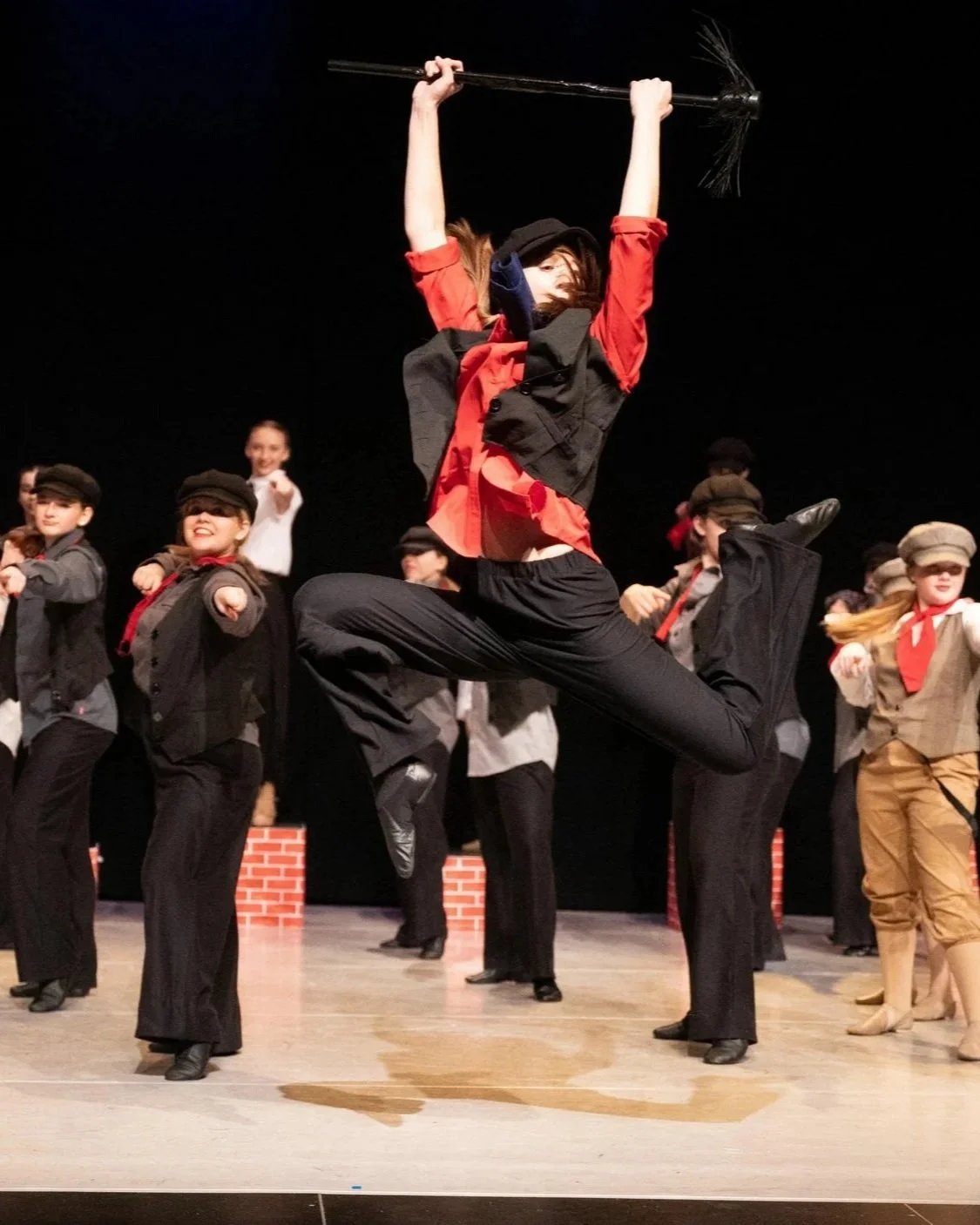 A group of children performing a theatrical dance on stage, with one girl jumping in the air holding a broomstick above her head, dressed in a red and black costume, while others wear vintage-style outfits with hats.