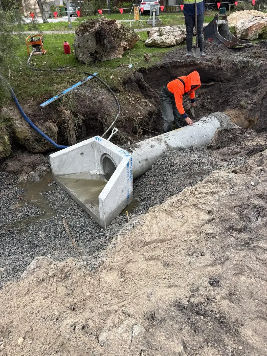 Workers installing a large RCP Stormwater pipe and Precast Headwall in an excavation site with trees, rocks, and construction equipment nearby.