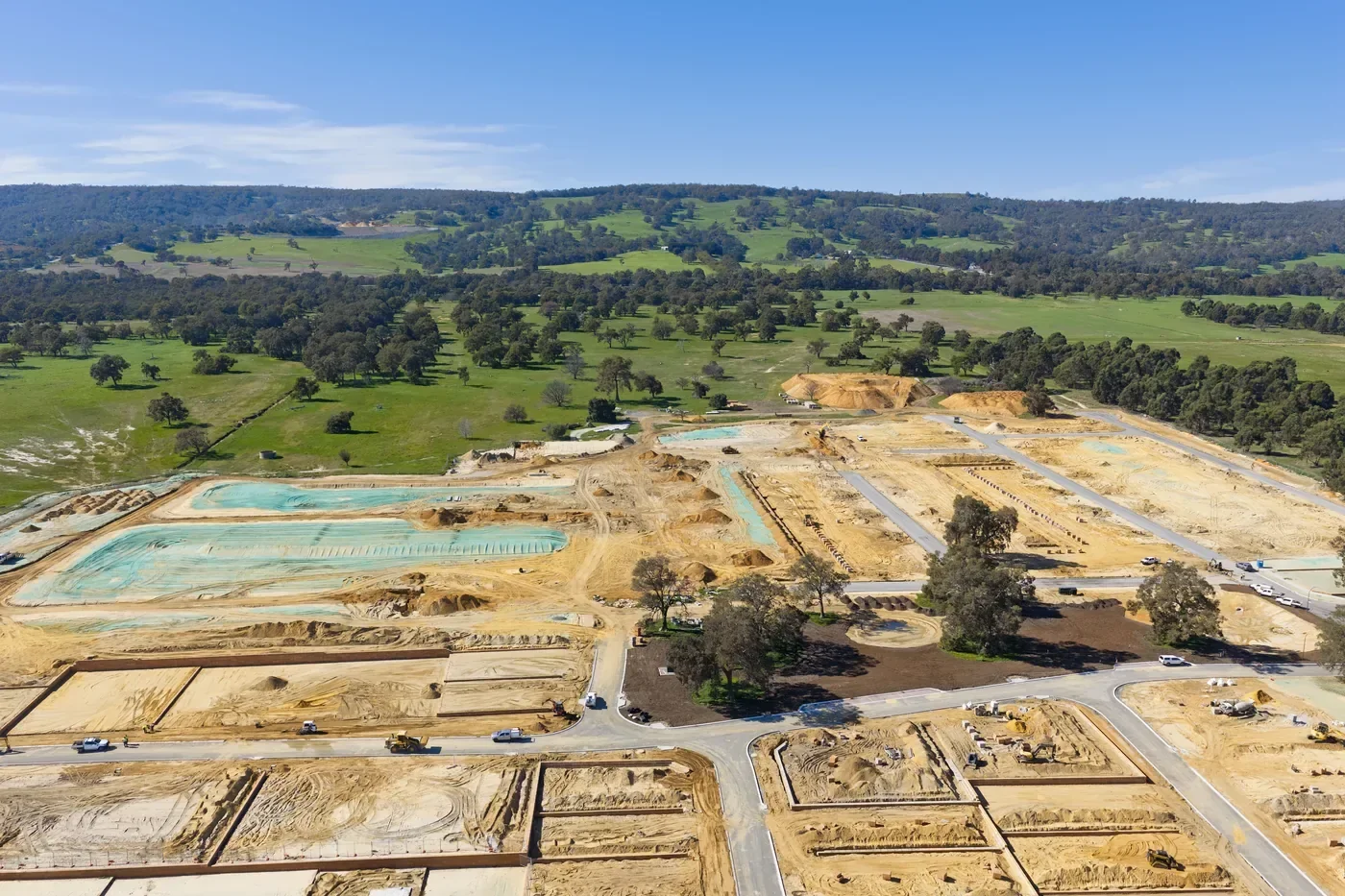 Aerial view of a construction site with excavation areas, roads, and some trees, set in a green hilly landscape under a blue sky.