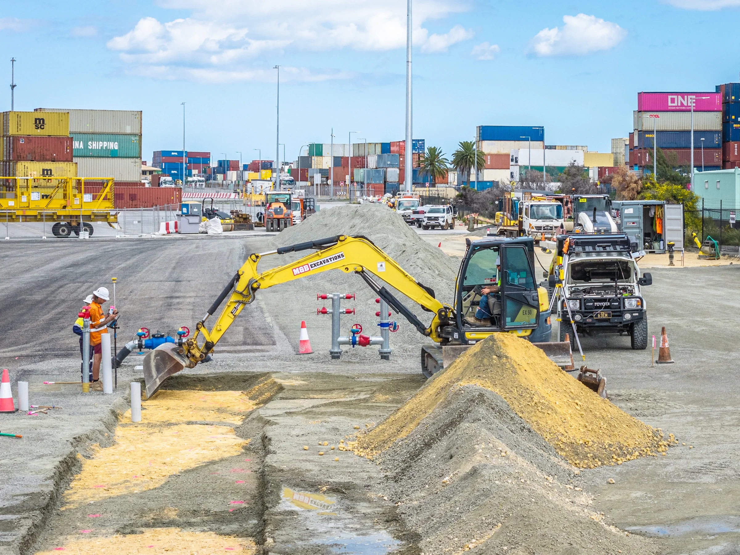 Construction workers and heavy machinery working on a port construction project with numerous shipping containers in the background under a partly cloudy sky.