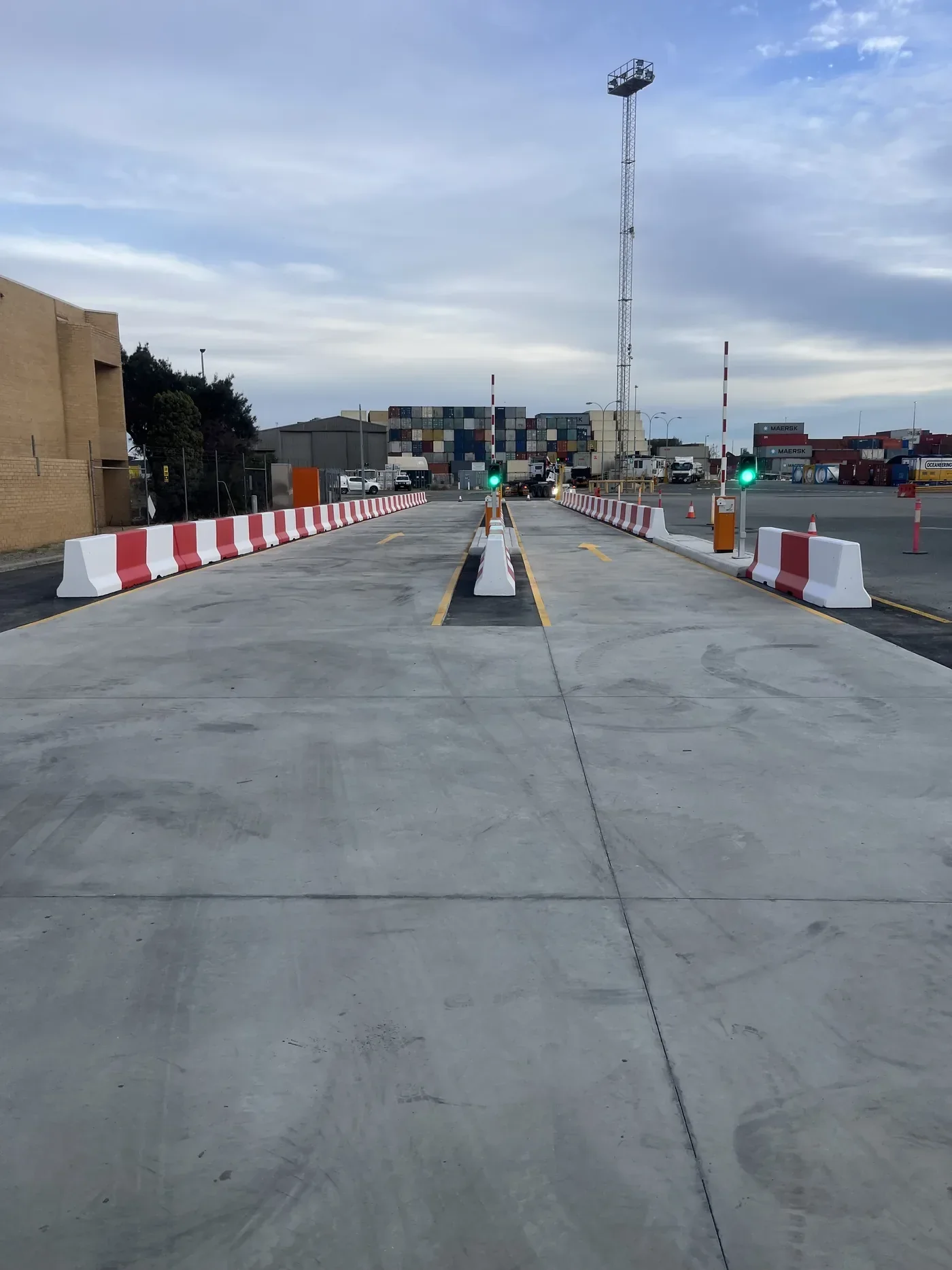 A completed weighbridge in an operating Port with shipping containers, crossing barriers, and traffic lights under a cloudy sky.