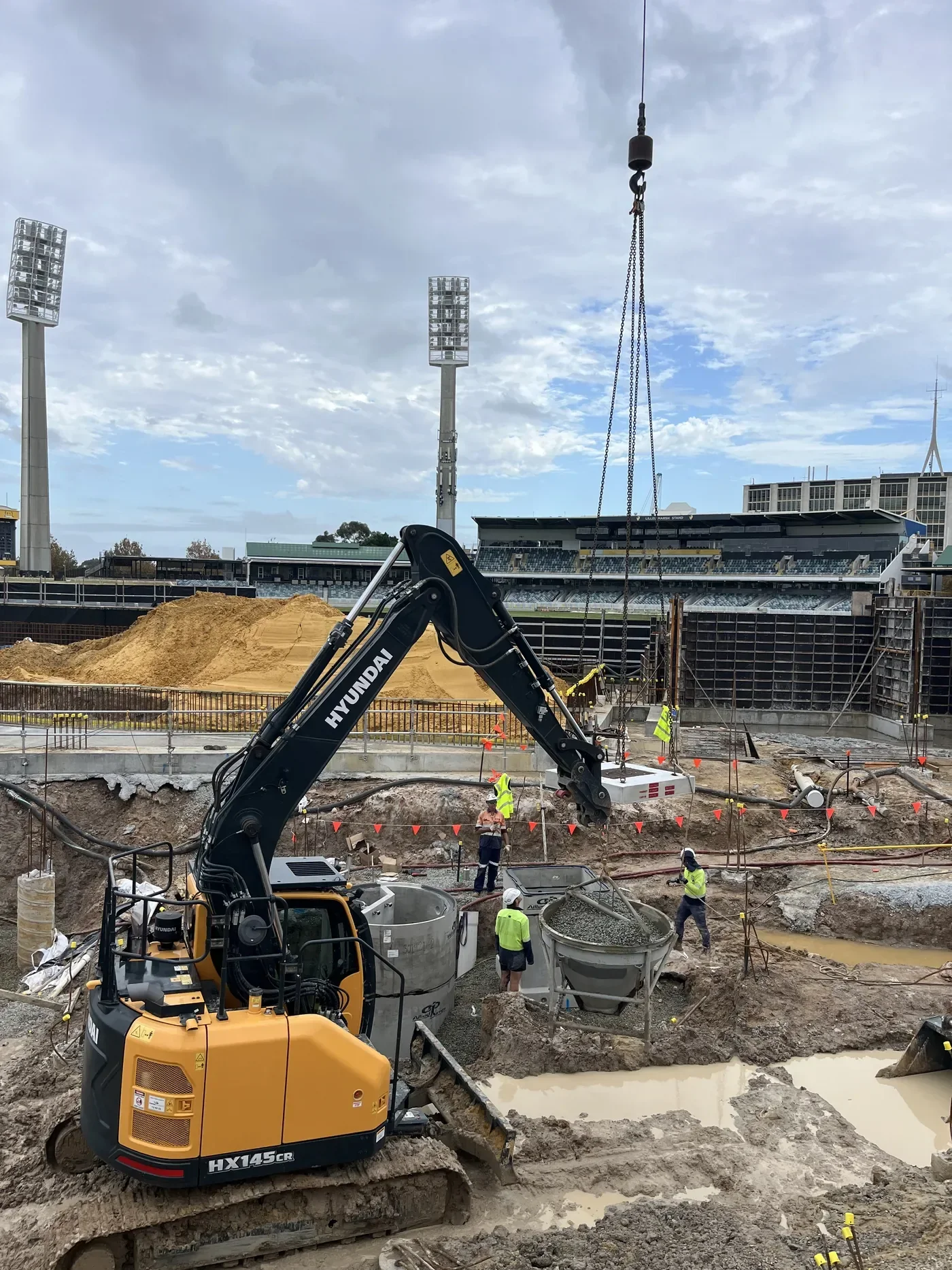 Construction site at the WACA Cricket ground in Western Australia with excavator, workers, and large piles of dirt, stadium with floodlights, and cloudy sky in the background.