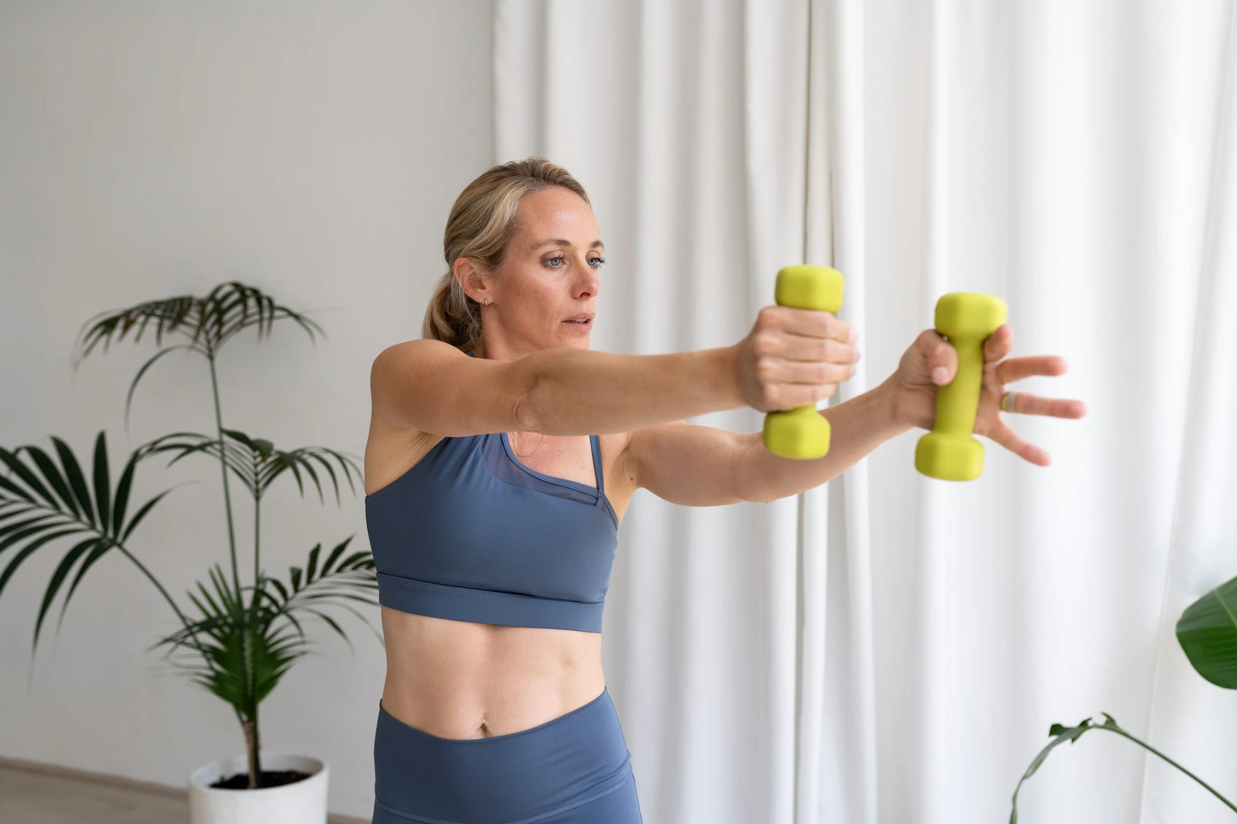 A woman in workout clothes lifting small green dumbbells indoors with plants in the background.