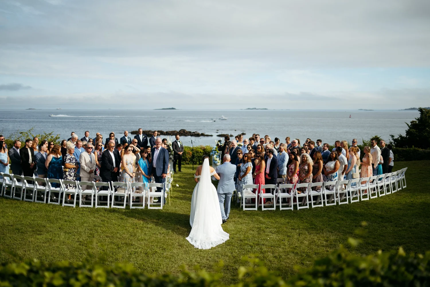 A bride walks down a lawn aisle by the ocean as guests stand on both sides