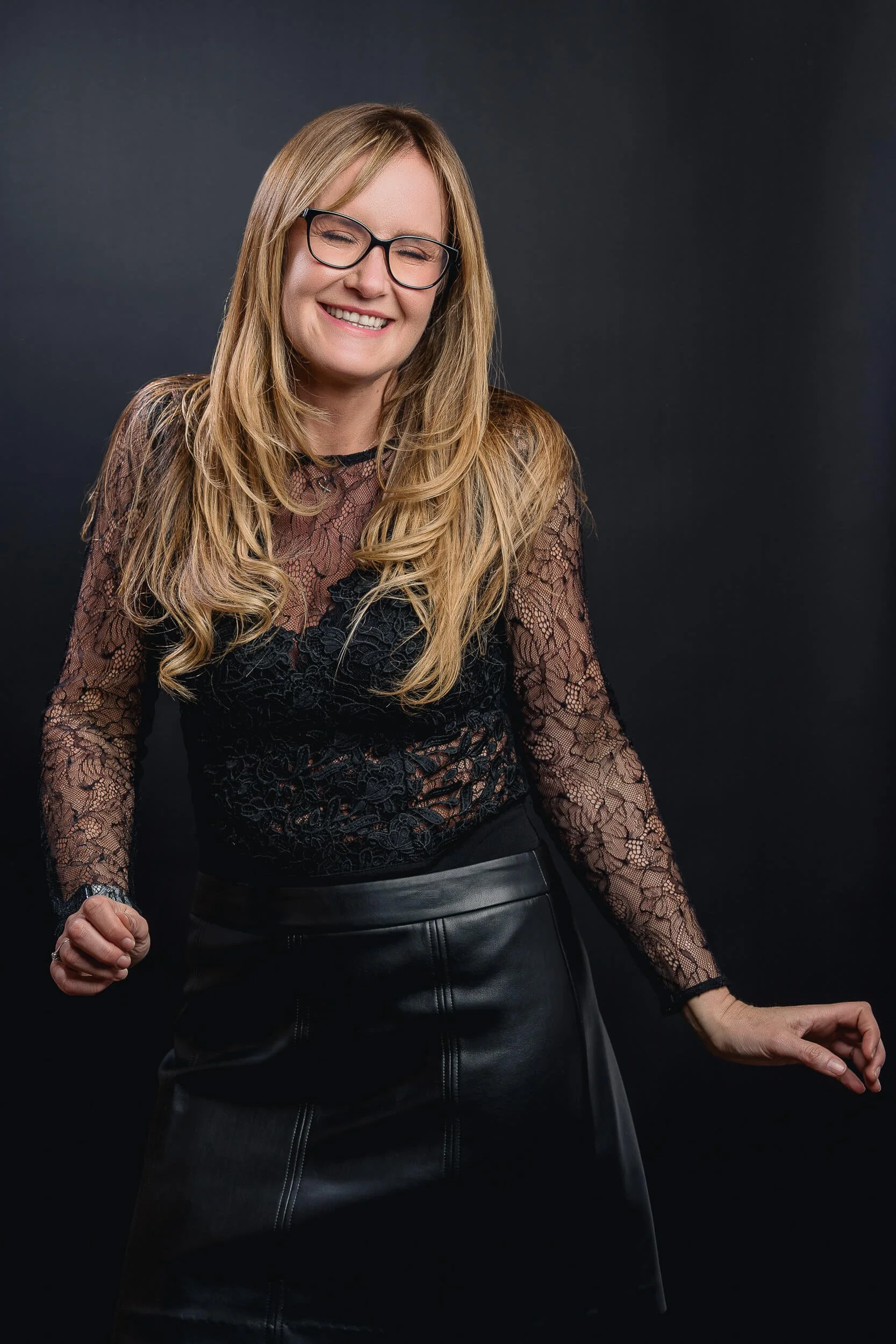 Studio portrait of a smiling woman with glasses wearing a black lace top against a dark background.